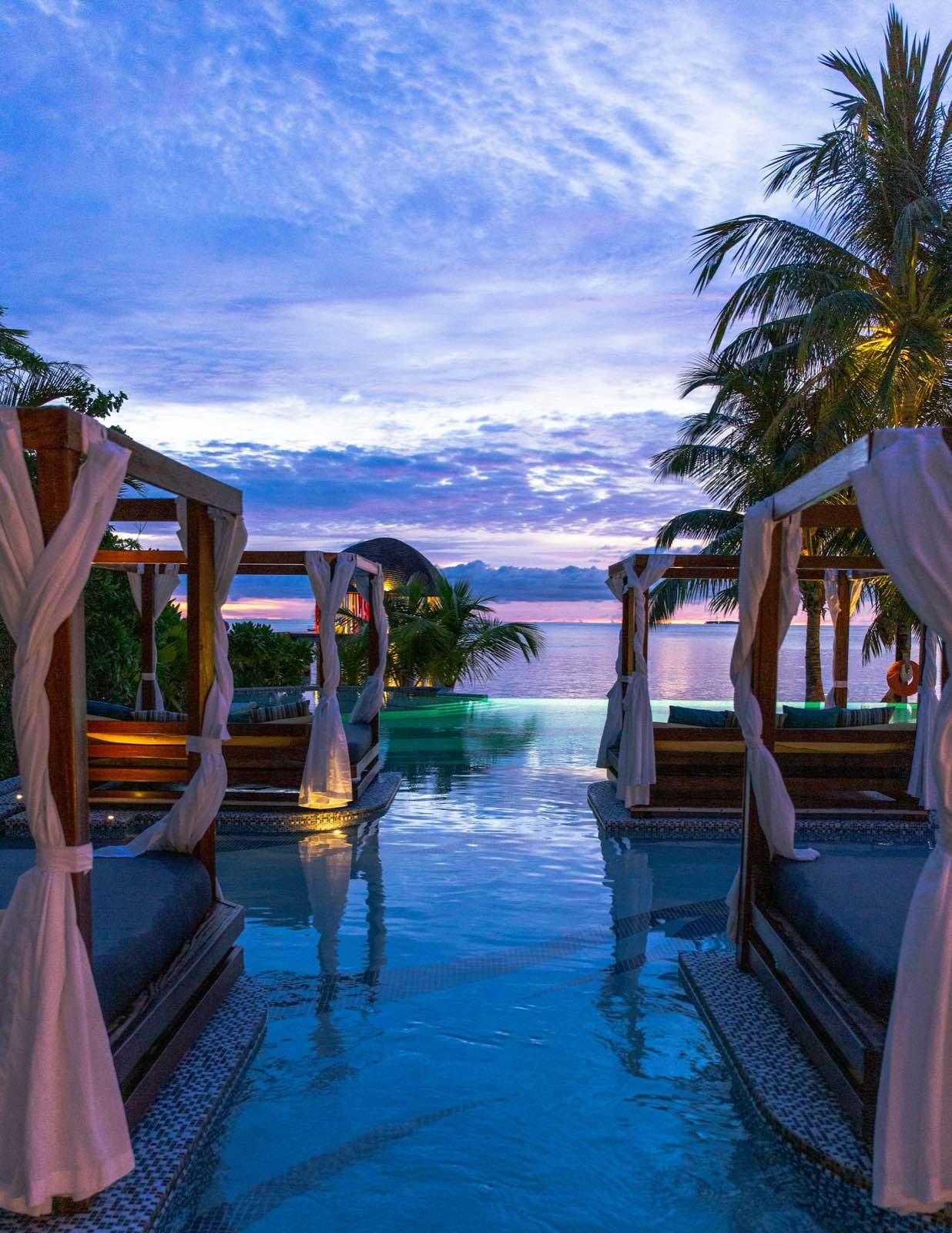 Camas balinesas con cortinas blancas a los lados de una piscina infinita, frente al mar al atardecer